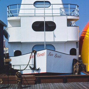 May include: A white boat with a red and white striped top and the words "Fun Boat" written on the side. The boat is docked at a pier with a wooden deck.