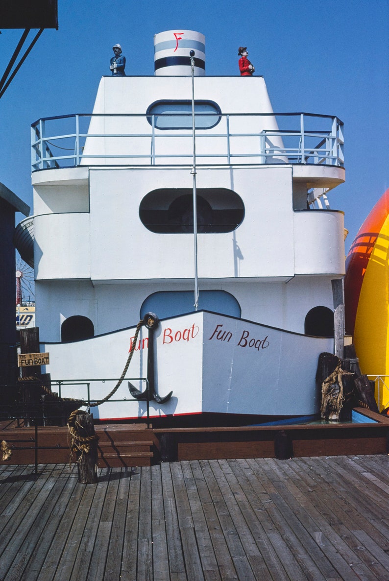 May include: A white boat with a red and white striped top and the words "Fun Boat" written on the side. The boat is docked at a pier with a wooden deck.