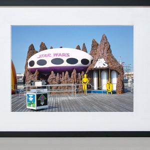 May include: A vintage Star Wars themed amusement park ride with a purple and white spaceship, a brown rock formation, and two yellow figures. The ride is on a wooden boardwalk.