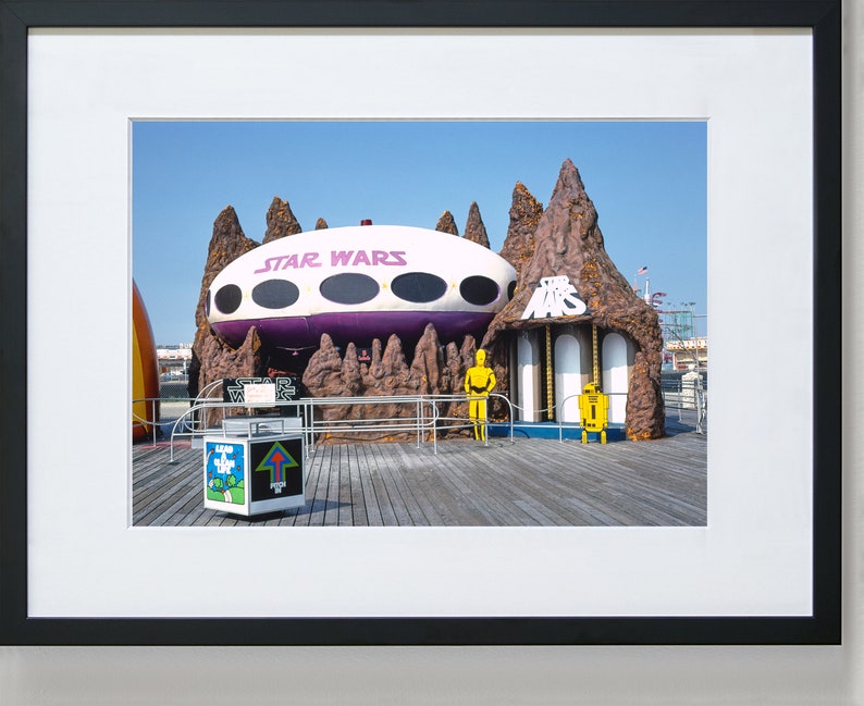 May include: A vintage Star Wars themed amusement park ride with a purple and white spaceship, a brown rock formation, and two yellow figures. The ride is on a wooden boardwalk.
