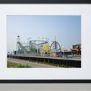 Op de afbeelding: Een ingelijste foto van een pretpark met een achtbaan en andere attracties. De achtbaan is groen en zilver, met de woorden "Supersonic Roller Coaster" zichtbaar. De lucht is helderblauw.