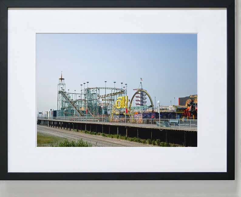 Op de afbeelding: Een ingelijste foto van een pretpark met een achtbaan en andere attracties. De achtbaan is groen en zilver, met de woorden "Supersonic Roller Coaster" zichtbaar. De lucht is helderblauw.