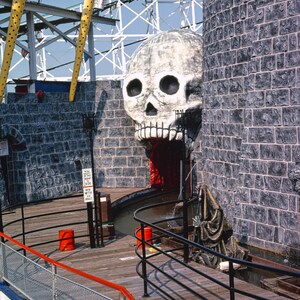 Puede incluir: Una gran escultura de calavera blanca con una boca roja que gotea es la entrada a un paseo oscuro en un parque de atracciones. La calavera est&aacute; situada sobre un fondo de muros de piedra gris. Una figura negra con una t&uacute;nica con capucha se encuentra frente a la calavera. Una pasarela de madera con barandillas negras conduce a la entrada del paseo. Un cartel indica "Debes medir al menos 48 pulgadas para montar. Un adulto debe acompa&ntilde;ar a cada ni&ntilde;o menor de 48 pulgadas."