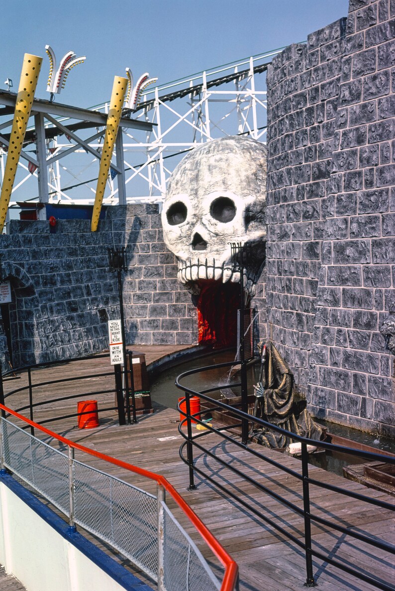 Puede incluir: Una gran escultura de calavera blanca con una boca roja que gotea es la entrada a un paseo oscuro en un parque de atracciones. La calavera est&aacute; situada sobre un fondo de muros de piedra gris. Una figura negra con una t&uacute;nica con capucha se encuentra frente a la calavera. Una pasarela de madera con barandillas negras conduce a la entrada del paseo. Un cartel indica "Debes medir al menos 48 pulgadas para montar. Un adulto debe acompa&ntilde;ar a cada ni&ntilde;o menor de 48 pulgadas."