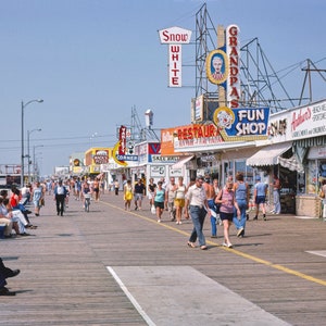Peut inclure: Une promenade anim&eacute;e avec des boutiques et des restaurants. Les boutiques ont des enseignes color&eacute;es avec des noms comme "Snow White", "Grandpa's" et "Fun Shop". Des gens se prom&egrave;nent sur la promenade, certains sont assis sur des bancs.