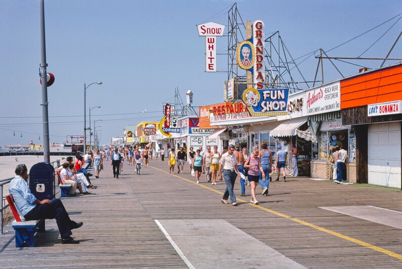 Peut inclure: Une promenade anim&eacute;e avec des boutiques et des restaurants. Les boutiques ont des enseignes color&eacute;es avec des noms comme "Snow White", "Grandpa's" et "Fun Shop". Des gens se prom&egrave;nent sur la promenade, certains sont assis sur des bancs.