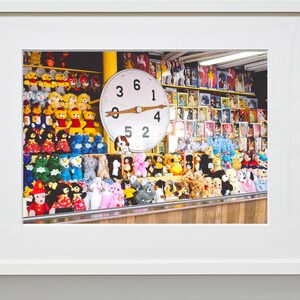 以下が含まれることがあります： 黒い数字と木製のポインターが付いた大きな白い時計の顔を持つカーニバルゲームのブース。ブースには、クマ、カエル、犬など、カラフルなぬいぐるみがぎっしり詰まっています。