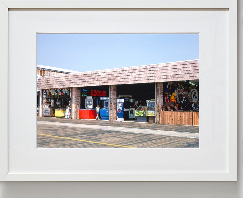 Pode incluir: Uma loja de arcade bem iluminada com uma placa vermelha que diz "ARCADE" e uma variedade de jogos de arcade vis&iacute;veis atrav&eacute;s das janelas. A loja est&aacute; localizada em um cal&ccedil;ad&atilde;o de madeira.