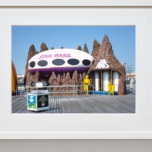 May include: A vintage photograph of a Star Wars themed amusement park ride. The ride features a purple and white spaceship with the words "Star Wars" on the side. The spaceship is partially obscured by a brown rock formation with the words "Star Wars" on the front. Two yellow robot figures stand in front of the ride.