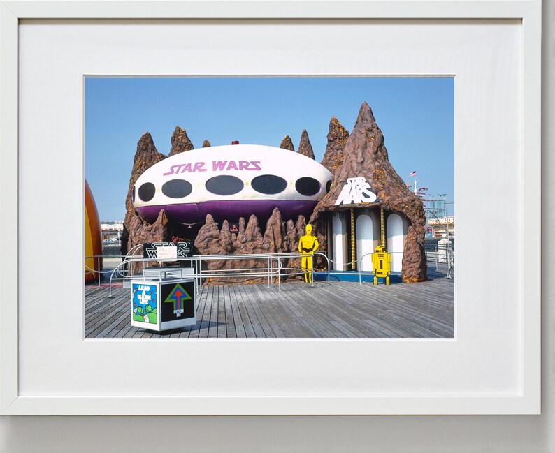 May include: A vintage photograph of a Star Wars themed amusement park ride. The ride features a purple and white spaceship with the words "Star Wars" on the side. The spaceship is partially obscured by a brown rock formation with the words "Star Wars" on the front. Two yellow robot figures stand in front of the ride.