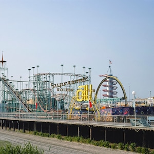 Op de afbeelding: Een pier van een pretpark met een achtbaan, een toren en andere attracties. De achtbaan is voorzien van het opschrift "Supersonic Roller Coaster". De pier is van hout en strekt zich uit in het water.