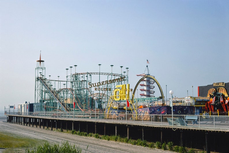 Op de afbeelding: Een pier van een pretpark met een achtbaan, een toren en andere attracties. De achtbaan is voorzien van het opschrift "Supersonic Roller Coaster". De pier is van hout en strekt zich uit in het water.