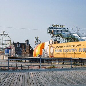 May include: A yellow and blue roller coaster with a sign that reads "Morey's Pier" in front of a yellow building with a sign that reads "Morey's Pier Presents... Bear Country Jubilee.  Annual Stage Show Direct From Orlando, Florida."