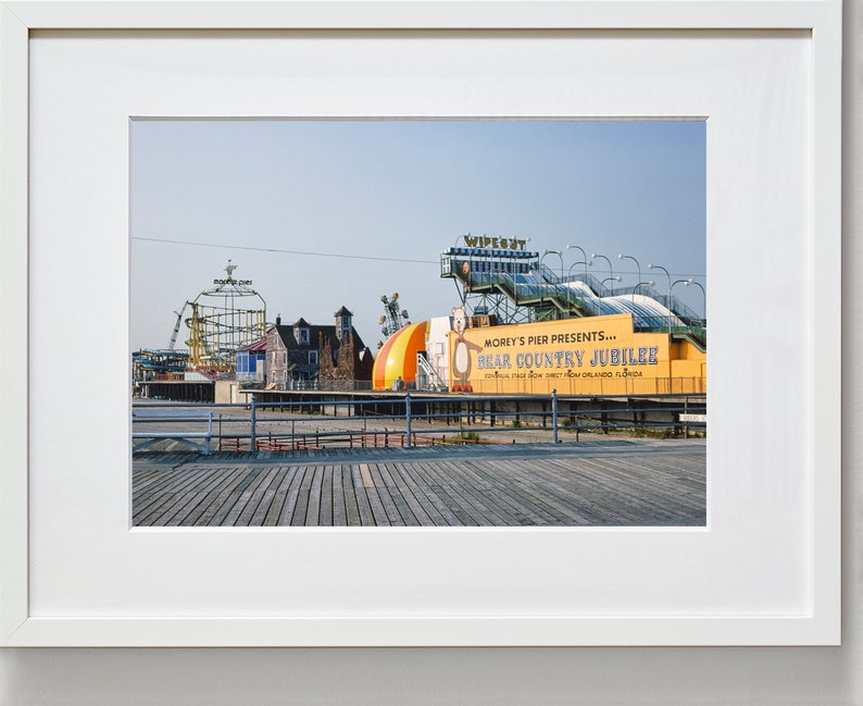 May include: A yellow and white sign with the text "Morey's Pier Presents... Bear Country Jubilee" on a boardwalk with a roller coaster and other amusement park rides in the background.
