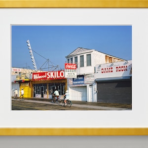 May include: A vintage photograph of a boardwalk scene with a bright blue sky. The image features a building with a sign that reads "Wildwood Skilo" and a sign that reads "Game Room". There are people riding bicycles in the foreground.