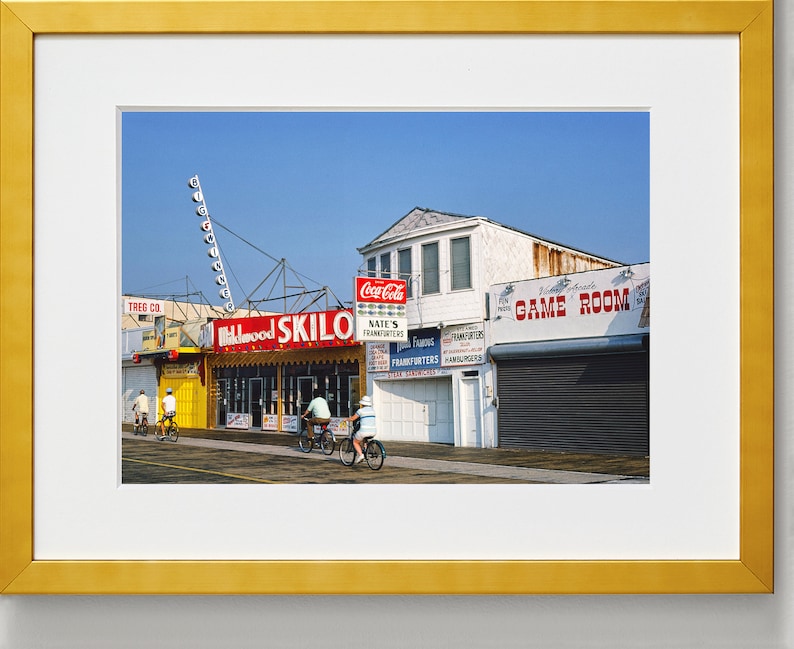 May include: A vintage photograph of a boardwalk scene with a bright blue sky. The image features a building with a sign that reads "Wildwood Skilo" and a sign that reads "Game Room". There are people riding bicycles in the foreground.