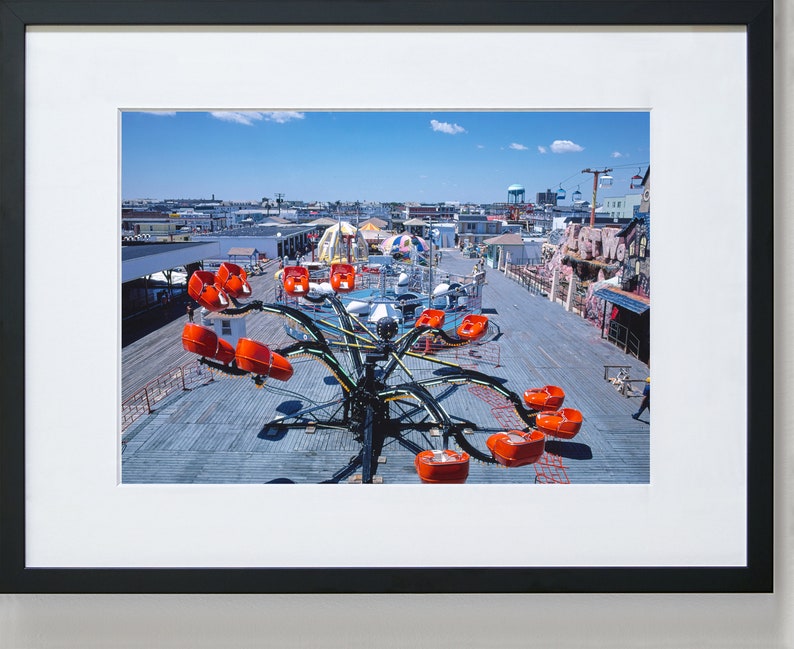 May include: A vintage photograph of a carnival ride with orange seats on a wooden boardwalk. The ride is a spinning octopus with long arms. The photo was taken from above, looking down on the ride.