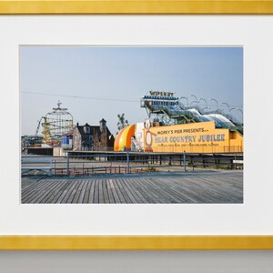 May include: A yellow and blue sign with the text "Morey's Pier Presents... Bear Country Jubilee" on a wooden boardwalk. The sign is in front of a colorful amusement park with a roller coaster and other rides.