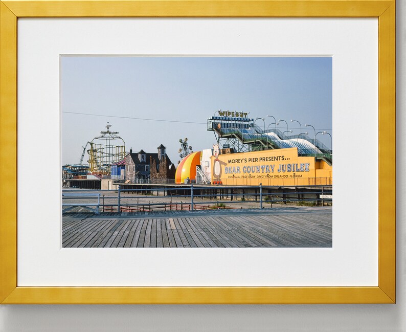 May include: A yellow and blue sign with the text "Morey's Pier Presents... Bear Country Jubilee" on a wooden boardwalk. The sign is in front of a colorful amusement park with a roller coaster and other rides.