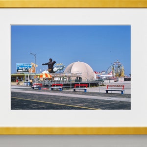 Op de afbeelding: Een vintage foto van een pretpark aan een boulevard met een groot, zwart gorillabeeld, een wit koepelvormig gebouw en een achtbaan op de achtergrond. De foto is gemaakt op een zonnige dag met een blauwe lucht.