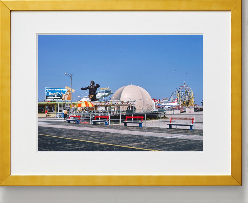 Op de afbeelding: Een vintage foto van een pretpark aan een boulevard met een groot, zwart gorillabeeld, een wit koepelvormig gebouw en een achtbaan op de achtergrond. De foto is gemaakt op een zonnige dag met een blauwe lucht.