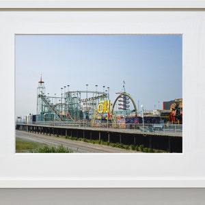 Op de afbeelding: Een ingelijste foto van een pretpark aan zee op een zonnige dag. De afbeelding toont een achtbaan met de woorden "Supersonic Roller Coaster" en andere attracties. De lucht is helderblauw en de sc&egrave;ne roept een gevoel van plezier en nostalgie op.