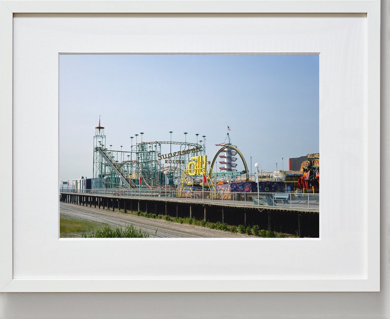 Op de afbeelding: Een ingelijste foto van een pretpark aan zee op een zonnige dag. De afbeelding toont een achtbaan met de woorden "Supersonic Roller Coaster" en andere attracties. De lucht is helderblauw en de sc&egrave;ne roept een gevoel van plezier en nostalgie op.