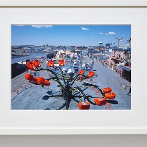 May include: A carnival ride with orange seats on a wooden boardwalk. The ride is a spinning octopus with long arms. The ride is in the middle of the boardwalk with other carnival rides and buildings in the background.