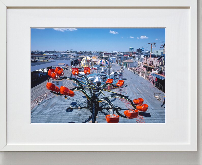 May include: A carnival ride with orange seats on a wooden boardwalk. The ride is a spinning octopus with long arms. The ride is in the middle of the boardwalk with other carnival rides and buildings in the background.