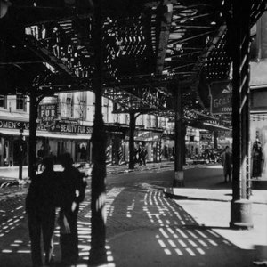 May include: A black and white photograph of a city street with a metal structure overhead. The structure casts shadows on the street below. There are signs for "Beauty Fur Shop" and "Gold Div" visible in the image.