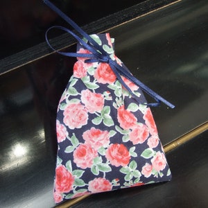Puede incluir: Una pequeña bolsa de cordón hecha de tela con un patrón floral. La bolsa es azul marino con rosas rosas y hojas verdes. La bolsa está atada con una cinta azul marino.