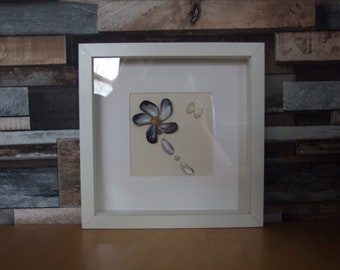 Könnte beinhalten: Ein gerahmtes Muschelkunstwerk mit einer Blume aus Muscheln auf weißem Hintergrund. Die Blume besteht aus weißen und violetten Muscheln und hat eine braune Mitte. Es gibt auch kleinere weiße Muscheln, die um die Blume herum verstreut sind.