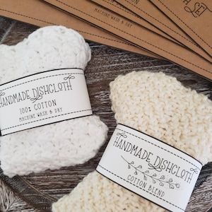 May include: Two handmade dishcloths, one white and one cream-colored, with labels that read "Handmade Dishcloth." The white dishcloth is 100% cotton and the cream one is a cotton blend. Both are machine washable and dryable.