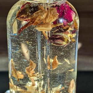 May include: A clear glass bottle with a black dropper top filled with a golden liquid and dried rose petals.
