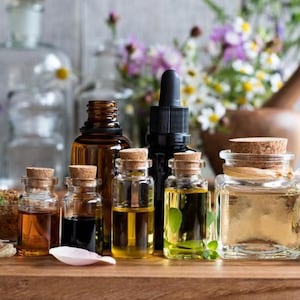 May include: A collection of various glass bottles containing essential oils, featuring amber, clear, and dark glass containers with cork stoppers and a black dropper cap. The bottles are arranged on a wooden surface with scattered flower petals.