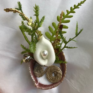 Könnte beinhalten: Eine weiße Calla-Lilie mit einer Perle in der Mitte, umgeben von grünem Laub und goldenem Glitzer, liegt in einer rosa Muschel mit einem braunen Garn-Akzent.