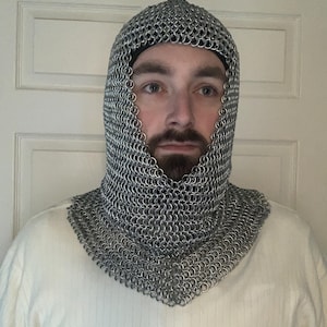 May include: A silver chainmail coif, a hood and neck covering made of interlocking metal rings. The coif is worn over a white long-sleeved shirt. The background is a white door.
