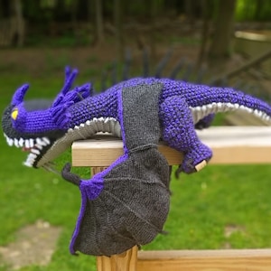 May include: A handmade, purple and gray crocheted dragon toy with large wings. The dragon has a detailed head with yellow eyes and white teeth. The wings are a mix of gray and purple. The dragon is resting on a wooden surface.