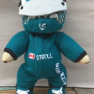 Puede incluir: Muñeca de punto a mano de color verde azulado que lleva una visera blanca y verde azulado con el texto "ASTON MARTIN". El atuendo verde azulado de la muñeca presenta "STROLL" y una bandera canadiense, con "ASTON MARTIN" en la pierna. La muñeca tiene zapatillas blancas con detalles en verde azulado.