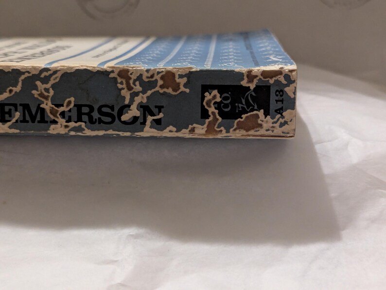 Op de afbeelding: Close-up van een boekrug met de naam "EMERSON" in zwarte letters. De rug is blauw met witte verticale lijnen en vertoont tekenen van slijtage. De rand van het boek heeft een logo en het nummer "A13".