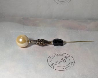 May include: A decorative pin with a large, off-white pearl, a silver-toned, ornate design, and a black bead. The pin is on a white background.