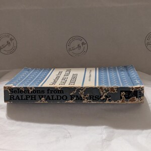 Op de afbeelding: Een vintage boek getiteld "Selections from Ralph Waldo Emerson" met een lichtblauwe kaft en witte tekst. De rug en randen van het boek vertonen tekenen van slijtage. De achtergrond is een wit, getextureerd oppervlak.