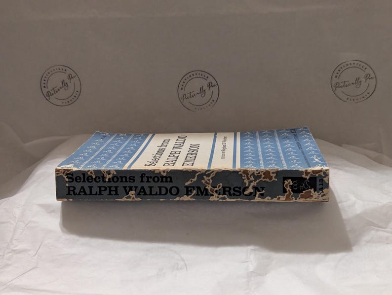 Op de afbeelding: Een vintage boek getiteld "Selections from Ralph Waldo Emerson" met een lichtblauwe kaft en witte tekst. De rug en randen van het boek vertonen tekenen van slijtage. De achtergrond is een wit, getextureerd oppervlak.