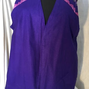 May include: A sleeveless purple vest with a deep V-neck. The shoulders are accented with a pink band and a decorative patterned fabric. The vest is displayed on a black mannequin.