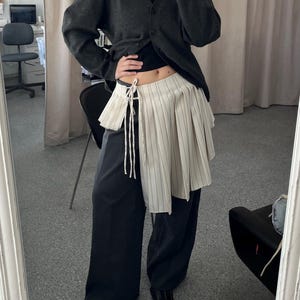 Könnte beinhalten: Dunkelgraue Strickjacke mit V-Ausschnitt, kombiniert mit einem schwarzen Oberteil und einer schwarzen Hose mit weitem Bein. Ein cremefarbener, plissierter Rock mit vertikalen Streifen ist über der Hose geschichtet, mit weißen Schnüren gebunden. Schwarze Schuhe vervollständigen das Outfit.