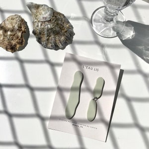 Puede incluir: Un par de pendientes de color verde claro con forma abstracta y un conector plateado. Los pendientes se muestran en una tarjeta blanca con el texto "L'EAU LIS" y "Crafted by hand inspired by nature @leau_lis".