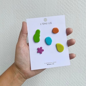 Pode incluir: Um conjunto de cinco alfinetes de argila coloridos num cartão de apresentação branco com a etiqueta L'EAU LIS e Crafted by hand @leau_lis. Os alfinetes incluem uma forma verde irregular, um pequeno círculo laranja, um oval azul, uma flor.
