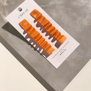 Op de afbeelding: Een paar oranje ruche-oorbellen op een witte kaart met de tekst "L'EAU LIS" en "Crafted by hand inspired by nature @leau_lis".