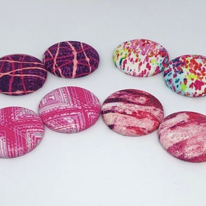 Puede incluir: Ocho botones redondos de tela con diferentes patrones rosas y morados. Los botones tienen un diámetro de aproximadamente 2,5 cm.
