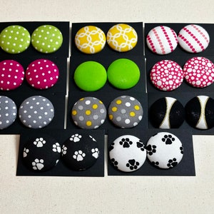 May include: A set of 10 pairs of fabric button earrings. The earrings are displayed on black cards. The earrings feature a variety of patterns, including polka dots, stripes, and paw prints. The colors include pink, green, yellow, gray, black, and white.